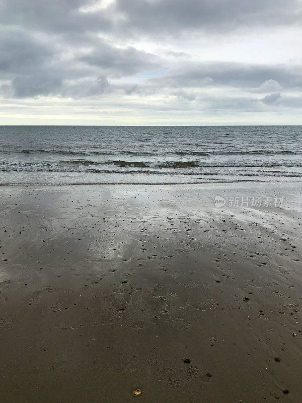 暴风雨的海滩日清晨，灰色的风暴云聚集在头顶上，黑暗的大海，温和的海浪，潮水拍打着海边的海岸线和海滩边潮湿的沙子，文字复制空间，沙滩上的脚印/狗的脚印，英国